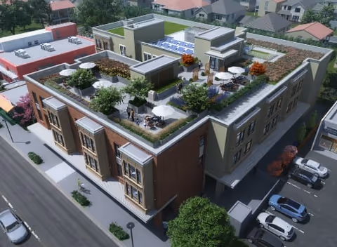 Aerial view of a modern senior living facility with a rooftop garden featuring seating areas, plants, and solar panels. The building is surrounded by streets, parked cars, and neighboring houses.