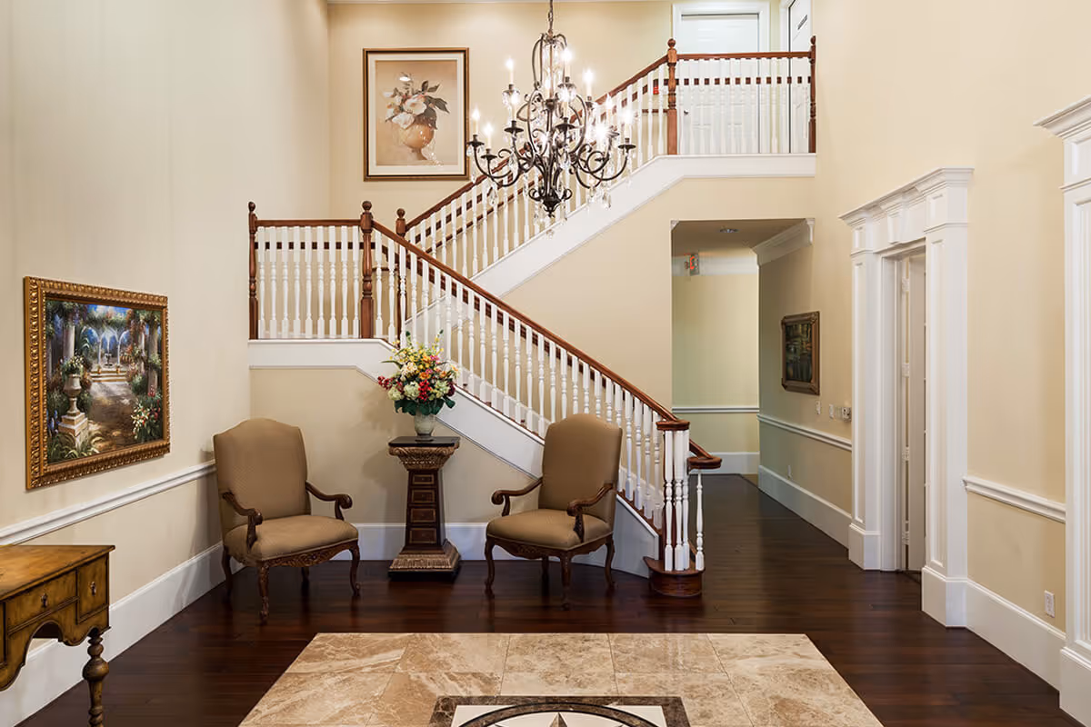 Interior view of a senior living facility featuring a grand staircase with wooden handrails and white balusters. Two upholstered armchairs flank a small pedestal table with a floral arrangement. The walls are painted beige and decorated with framed artwork. A chandelier hangs from the ceiling, illuminating the space with warm light. The floor is a combination of dark wood and beige tile with a decorative pattern.