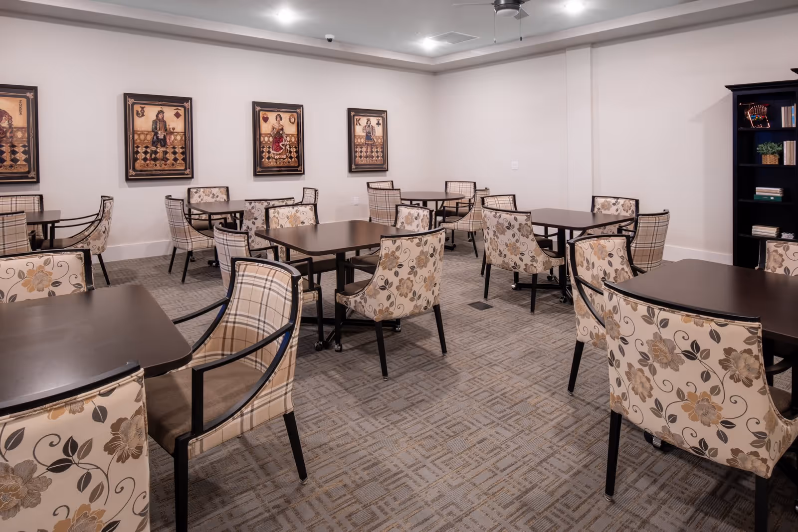 A dining room with several dark wood tables and upholstered chairs featuring floral and plaid patterns. The walls are light-colored and decorated with framed artwork depicting playing cards. There is a black bookshelf with books and decorative items on the right side of the room.