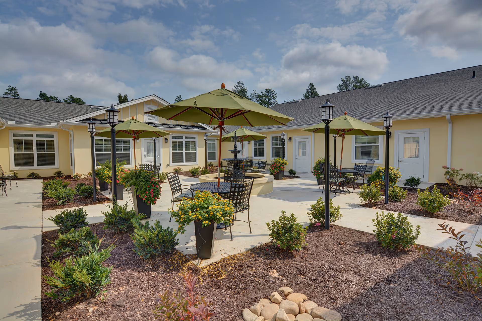 Outdoor courtyard area at Barclay House of Augusta featuring multiple green patio umbrellas over black metal tables and chairs, surrounded by landscaped garden beds with various shrubs and flowers. The courtyard is paved with concrete walkways and has a central water fountain. The building exterior is light yellow with white trim and multiple windows and doors.
