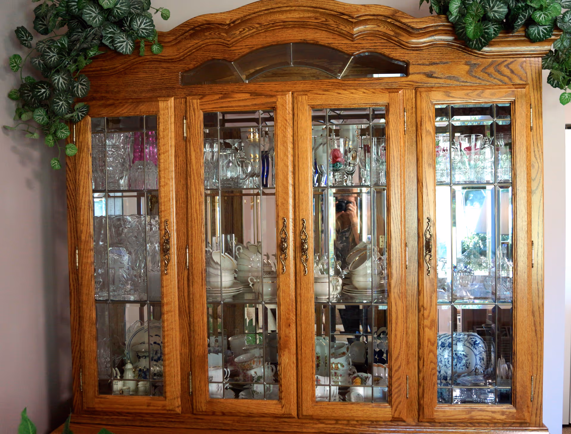 A wooden china cabinet with glass-paneled doors displaying various glassware, teacups, plates, and decorative dishes. There are green leafy plants placed on top of the cabinet.