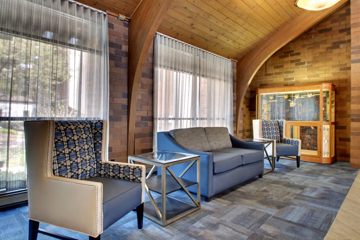 A cozy sitting area with two patterned armchairs and a blue sofa arranged around two glass-top side tables. The room features large windows with sheer curtains, exposed brick walls, and a wooden arched ceiling. A wooden display cabinet is visible in the background.