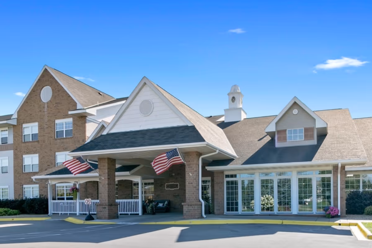 Front entrance of a multi-story senior living building with a covered porte-cochère, American flags, large windows and brick facade.