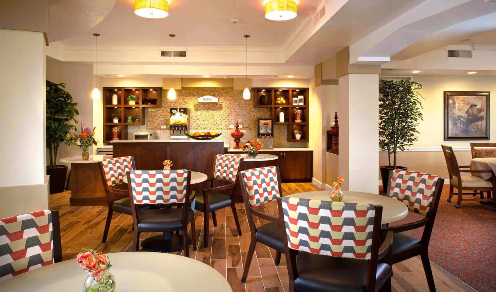 A bright and inviting dining area in an assisted living facility featuring round tables with patterned chairs, small flower vases on each table, wooden flooring, and a beverage station labeled 'Bistro' in the background with pendant lights hanging from the ceiling.