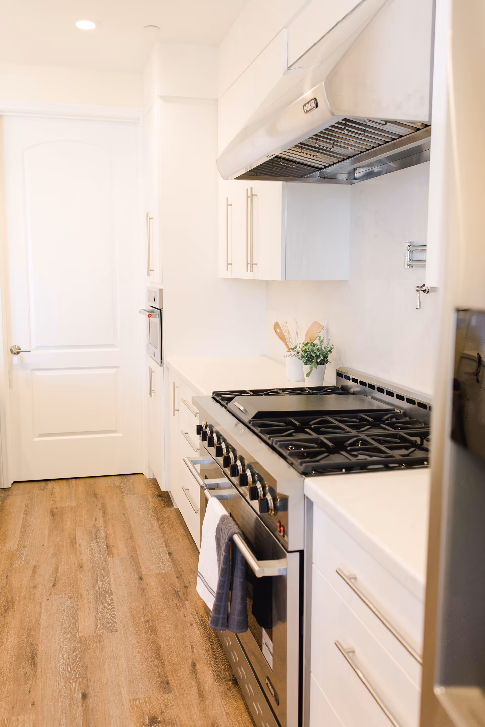 A modern kitchen with white cabinets, a stainless steel gas stove with multiple burners, a stainless steel range hood, and wooden flooring. There are kitchen utensils in a white container and a small green plant on the countertop. A white door is visible at the end of the kitchen.