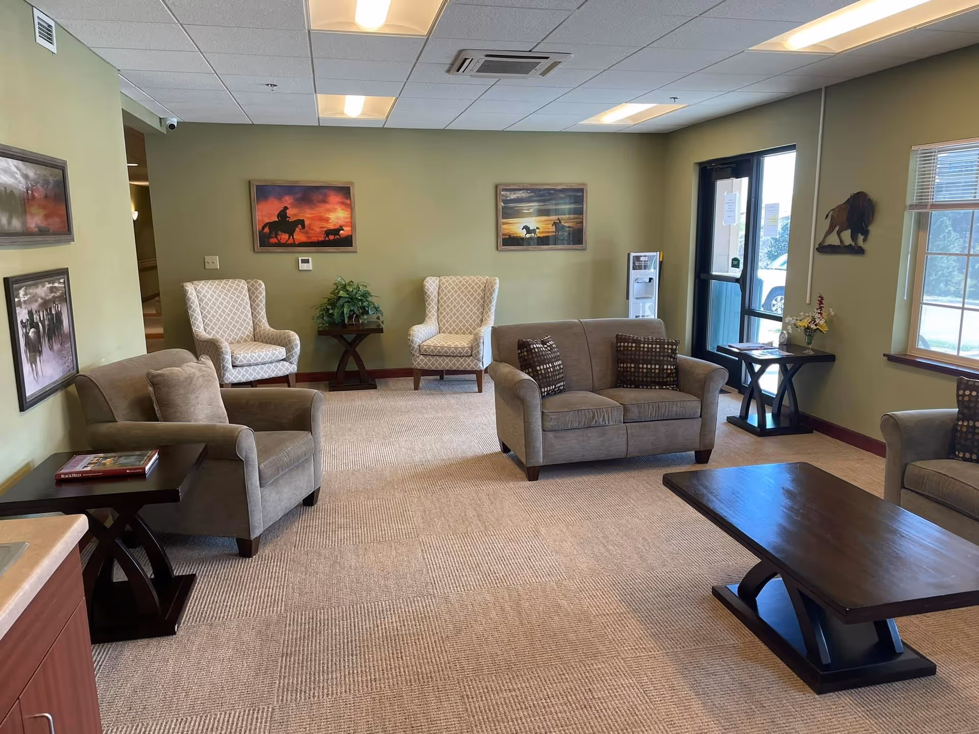 A cozy living room area with beige carpet and light green walls. The room features two patterned armchairs against the back wall, a loveseat with two patterned pillows, and two single armchairs. There are three framed pictures on the walls depicting horses and a cowboy, a wooden coffee table in the center, and two side tables with decorative items. A water dispenser is near the glass door, and a window lets in natural light.