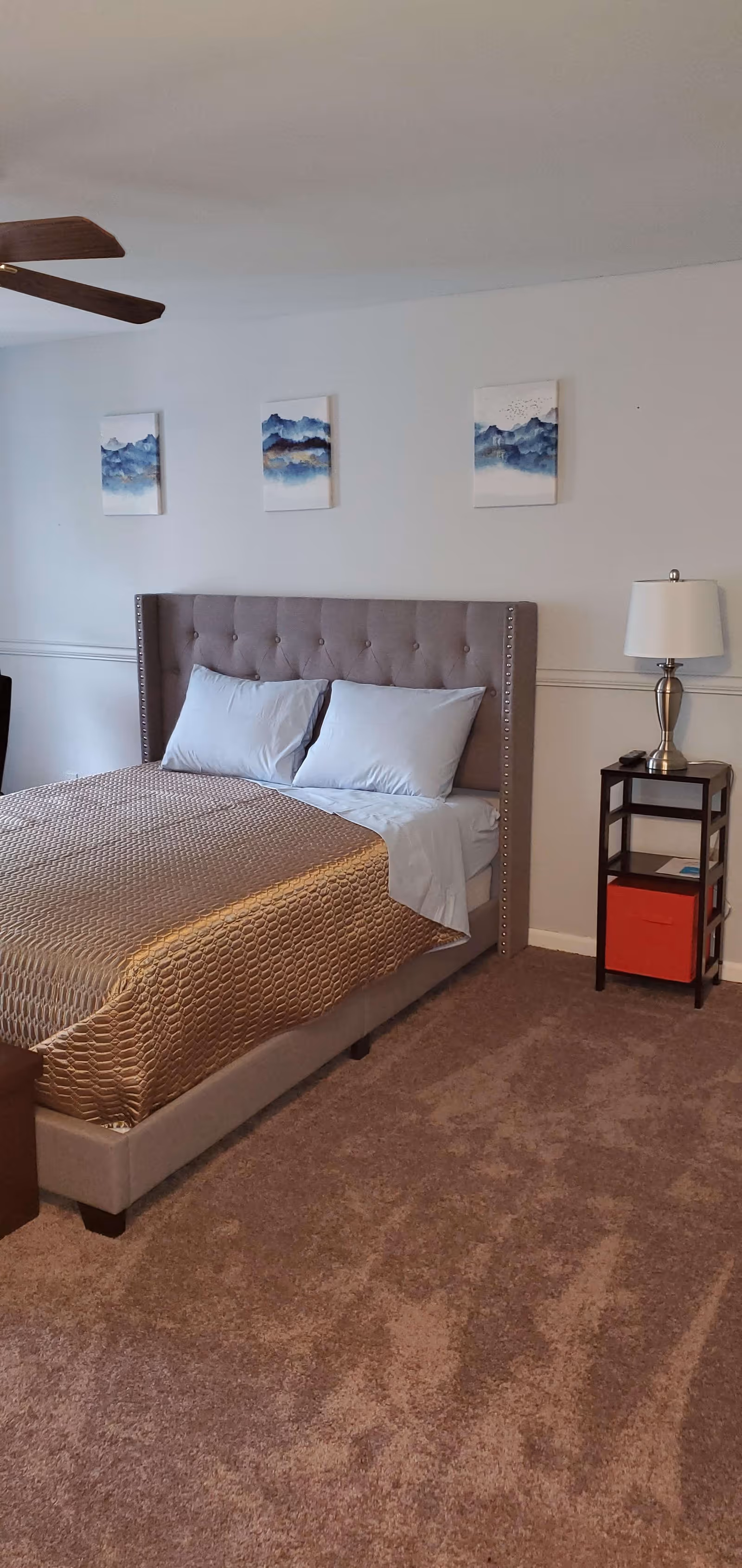 A bedroom with a beige tufted headboard bed made with light blue pillows and a gold textured bedspread. Above the bed are three framed paintings of blue and white abstract landscapes. To the right of the bed is a small dark wooden nightstand with a silver lamp, a remote control, and a red storage box on the lower shelf. The room has beige carpet and white walls.