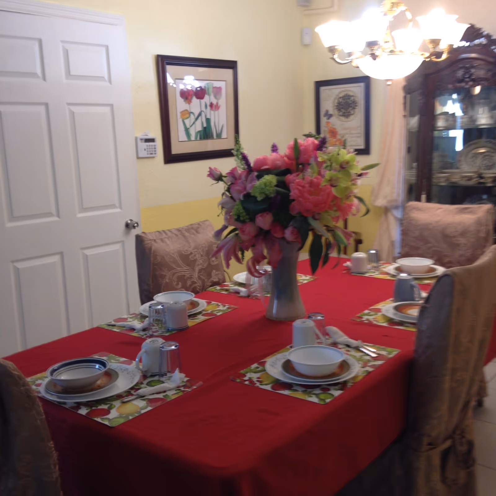 A dining room with a rectangular table covered with a red tablecloth, set with bowls, plates, cups, and utensils on colorful placemats. A large vase with a bouquet of pink and green flowers is in the center of the table. The room has cushioned chairs with patterned covers, a white door, framed floral artwork on the walls, and a glass cabinet displaying dishes. A chandelier hangs above the table providing light.