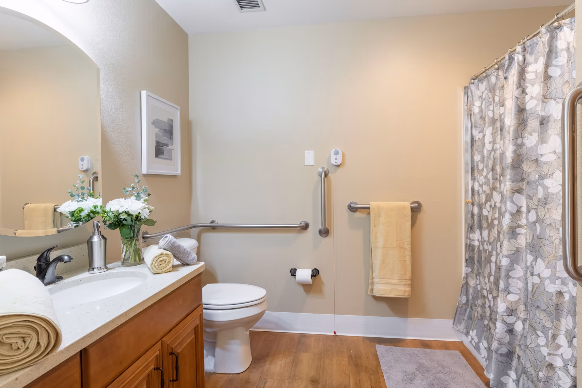 A clean and accessible bathroom with a toilet, a sink with a countertop holding rolled towels and a vase of white flowers, a large mirror, grab bars near the toilet and shower, a towel hanging on a bar, and a shower with a floral-patterned curtain.