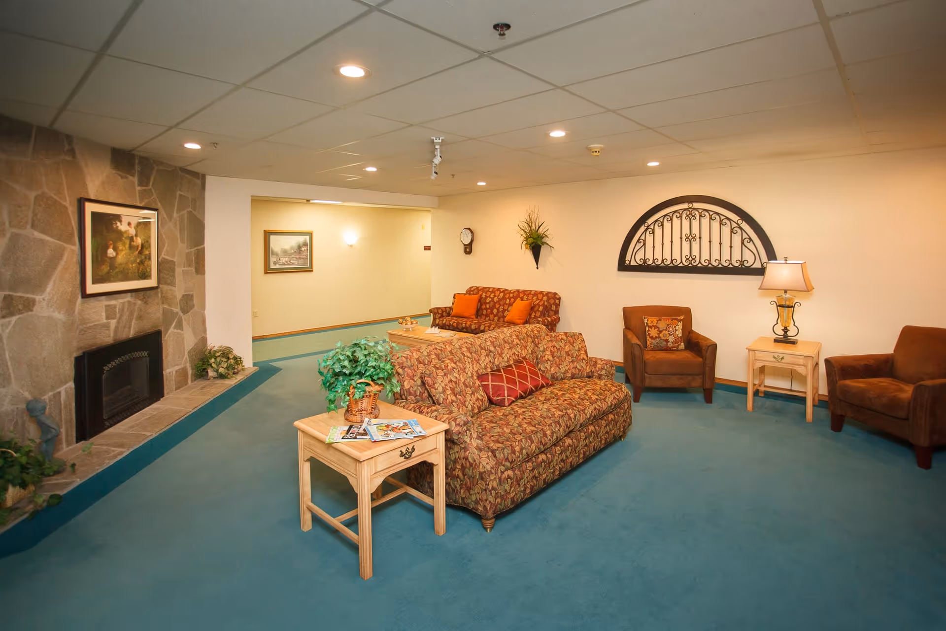A cozy living room area with teal carpet, two patterned sofas with orange and red pillows, two brown armchairs, and light wooden side tables. The room features a stone fireplace with a framed painting above it, a wall clock, a decorative wall-mounted plant, and a lamp on one of the side tables. The walls are light-colored, and the ceiling has recessed lighting.