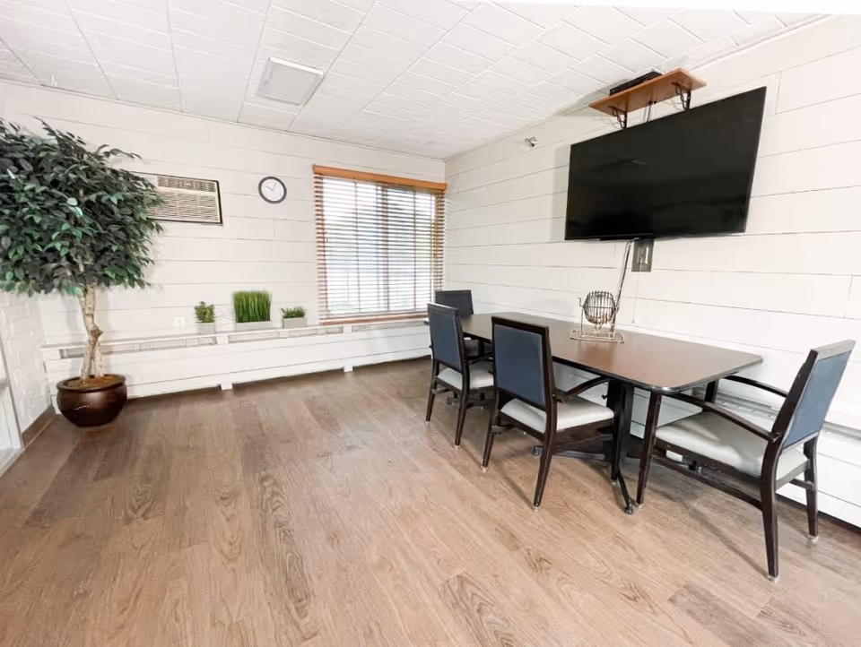 A small meeting or activity room with a rectangular table and four chairs. A large flat-screen TV is mounted on the white shiplap wall above the table. There is a window with wooden blinds, a wall clock, a framed picture, and a potted plant in the corner. The floor is wood, and the room has a clean, simple, and bright appearance.
