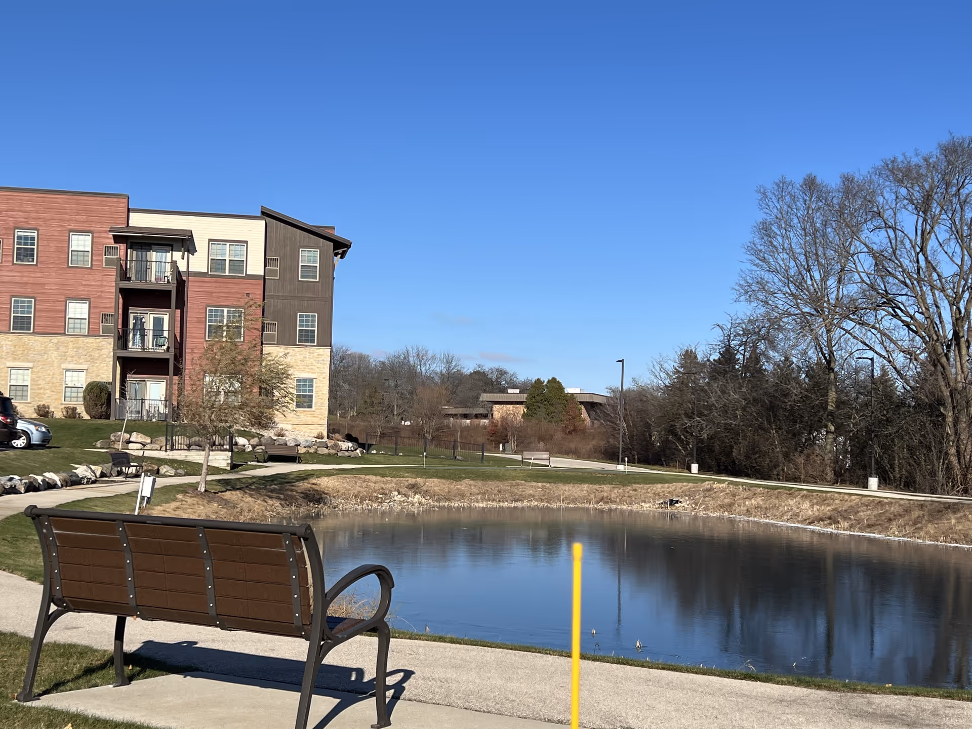 Outdoor scene at Hamilton House Senior Living showing a small pond with a walking path around it, a brown bench in the foreground, a multi-story residential building to the left, and trees in the background under a clear blue sky.