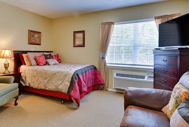A cozy bedroom with a bed dressed in beige and red bedding, a green armchair, a brown leather chair with a floral pillow, a wooden dresser with a TV on top, a window with beige curtains, and two framed pictures on the wall.