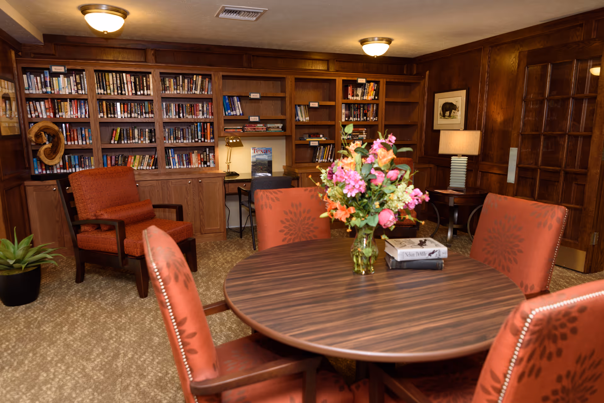 A cozy library room with wood-paneled walls and built-in bookshelves filled with books. There is a round wooden table in the center with a vase of colorful flowers and two books on it, surrounded by four upholstered chairs with a floral pattern. In the background, there is a comfortable armchair, a desk with a lamp, a framed picture on the wall, and a potted plant on the floor.