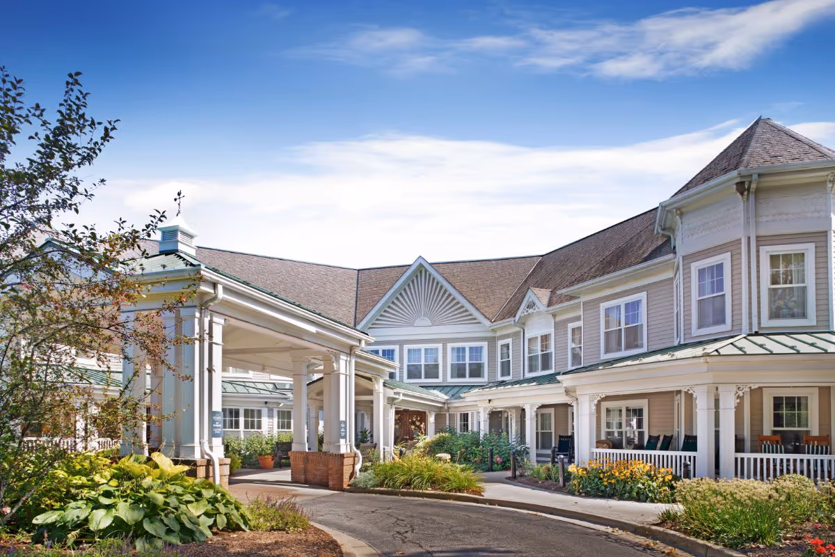 Front entrance of a two-story senior living building with a covered porte-cochere, wraparound porch, and landscaped gardens.