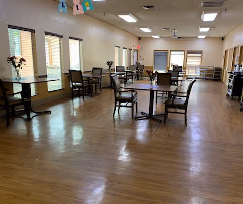 A spacious room with wooden flooring featuring multiple round tables and chairs arranged neatly. Each table has a small flower vase as decoration. The room has several windows with blinds allowing natural light to enter. The ceiling has fluorescent lights and a ceiling fan. The overall setting suggests a communal dining or gathering area.