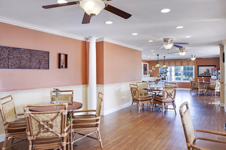 A bright dining area with multiple round tables and cushioned chairs arranged on a wooden floor. The walls are painted in a warm peach color with white trim, and there are ceiling fans and recessed lights overhead. Large windows allow natural light to fill the room, and decorative wall art is visible.