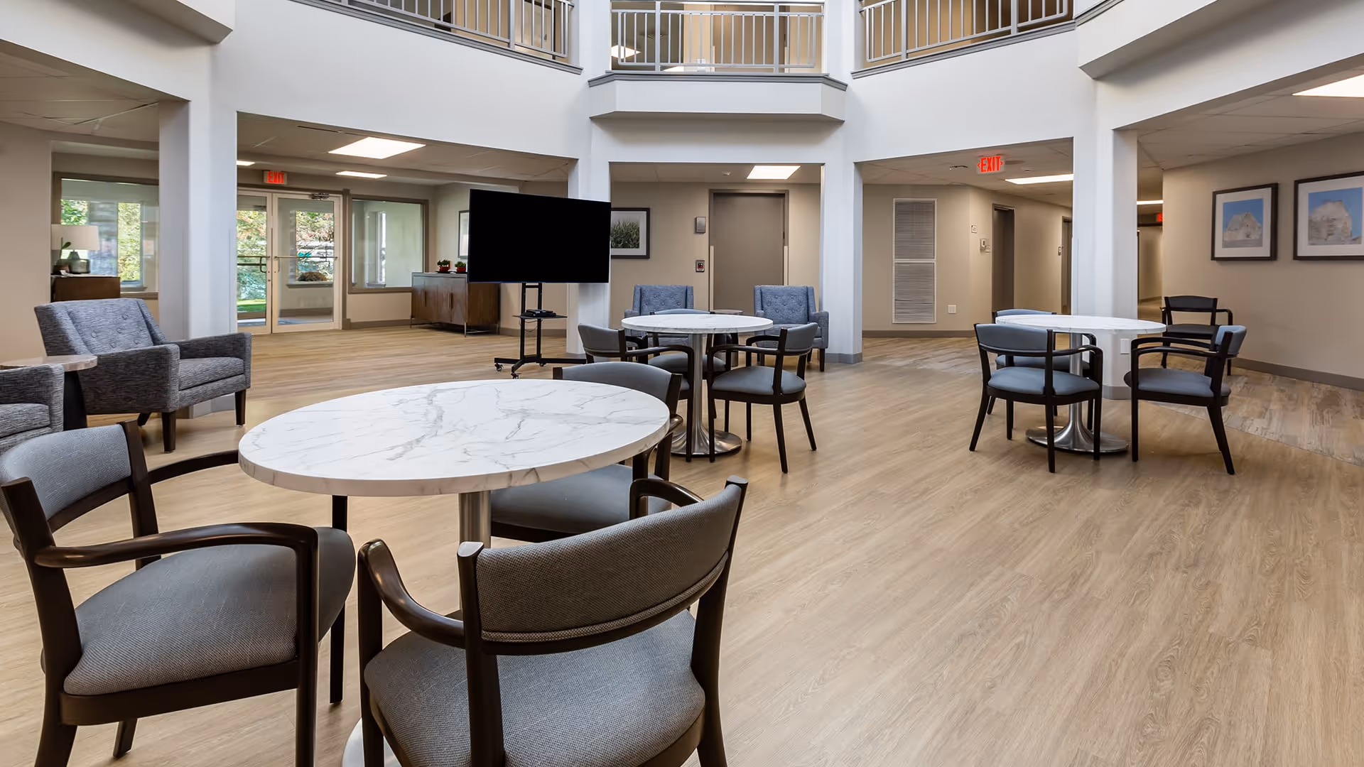 A spacious, well-lit common area with round marble-top tables and cushioned chairs arranged for seating. The room features light wood flooring, neutral-colored walls, and a high ceiling with a balcony railing above. There is a large flat-screen TV on a stand, several armchairs, framed artwork on the walls, and glass doors leading outside.