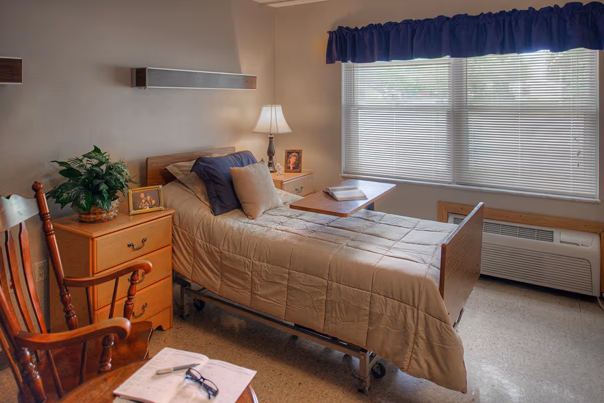 A single bed with beige bedding and multiple pillows in a room with a wooden rocking chair, a small wooden nightstand with a plant and framed photos, a table over the bed with an open book, a lamp on a side table, and a large window with blinds and a blue valance.
