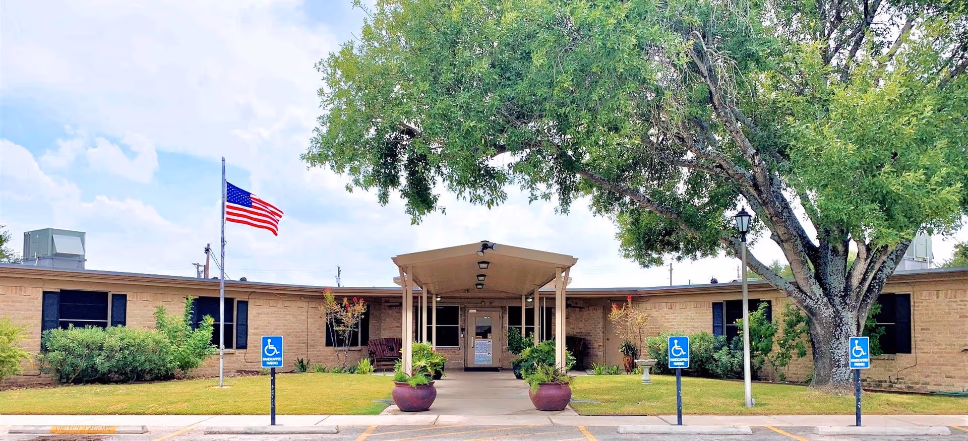 Front exterior view of Devine Health and Rehabilitation facility showing a single-story brick building with a covered entrance, an American flag on a flagpole, handicap parking signs, and a large tree on the right side.