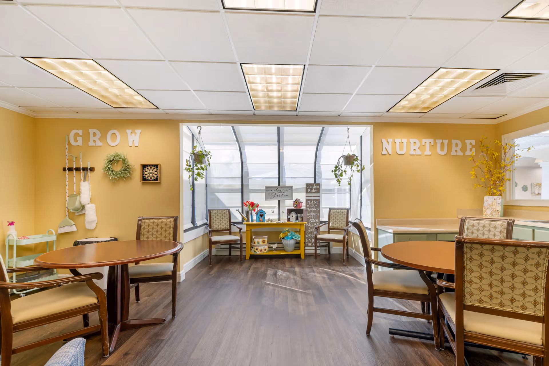 Bright communal activity room with round tables and chairs, plants, and decorative letters spelling 'GROW' and 'NURTURE' on yellow walls.