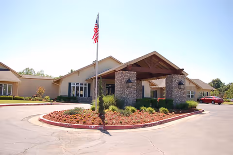Front exterior view of Cedar Ridge Senior Living facility with a covered entrance supported by stone pillars, an American flag on a flagpole, landscaped flower beds, and a driveway with a fire lane.