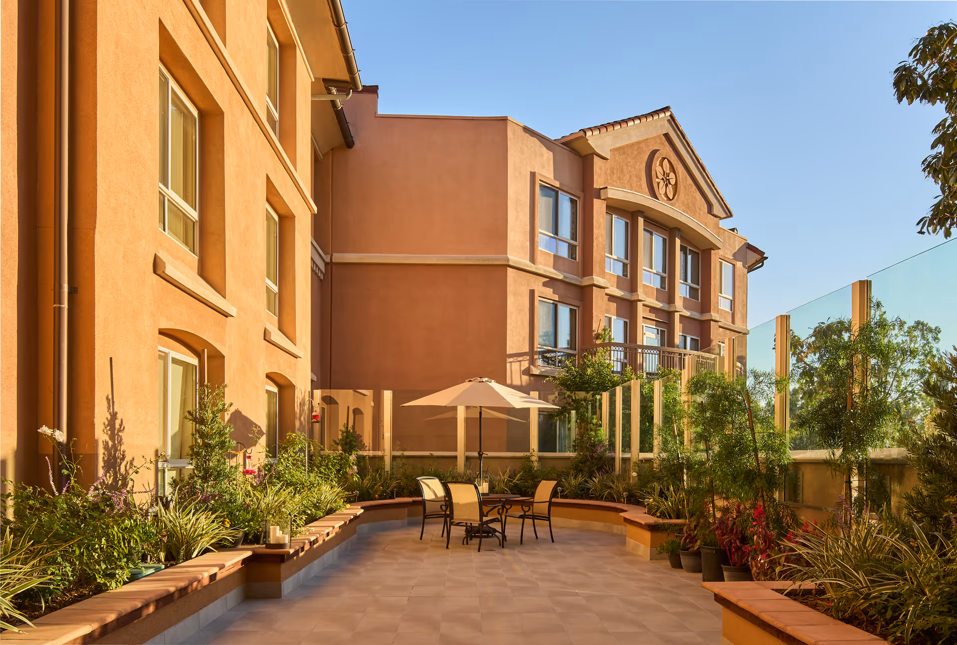 Outdoor patio area at MorningStar Senior Living of Pasadena with a round table, four chairs, and a large umbrella surrounded by plants and greenery. The patio is adjacent to a multi-story building with large windows and a clear blue sky overhead.