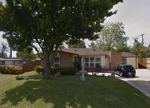 Front exterior view of a single-story assisted living facility named Mendoza #2 Assisted Living, featuring a well-maintained lawn, a large tree, a driveway with a parked black car, and a brick chimney.
