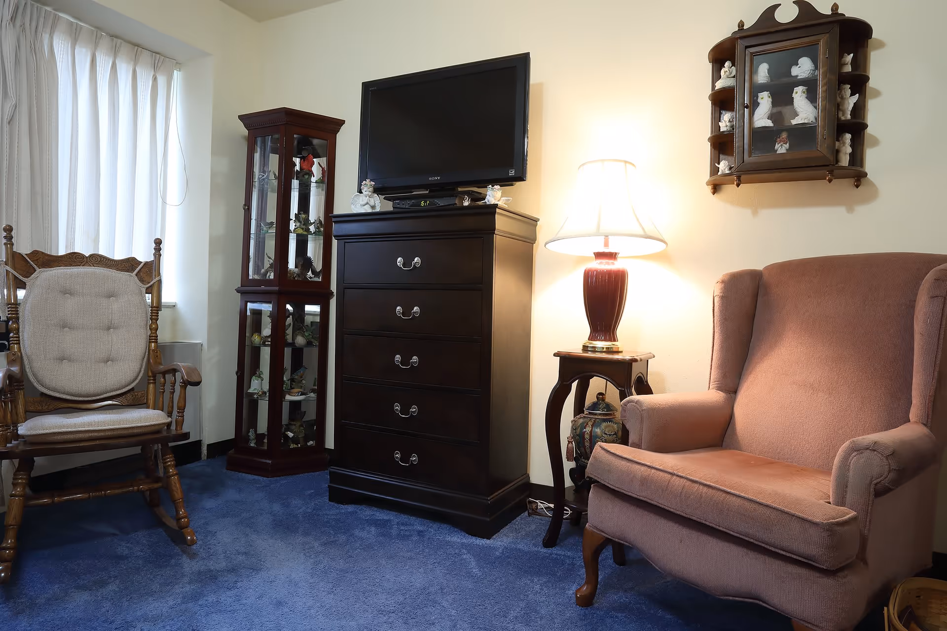 A cozy living room area with a wooden rocking chair with a cushion, a tall glass display cabinet filled with figurines, a dark wooden chest of drawers with a flat-screen TV on top, a small side table with a red lamp, and a pink upholstered armchair. The floor is covered with blue carpet and there is a window with white curtains.