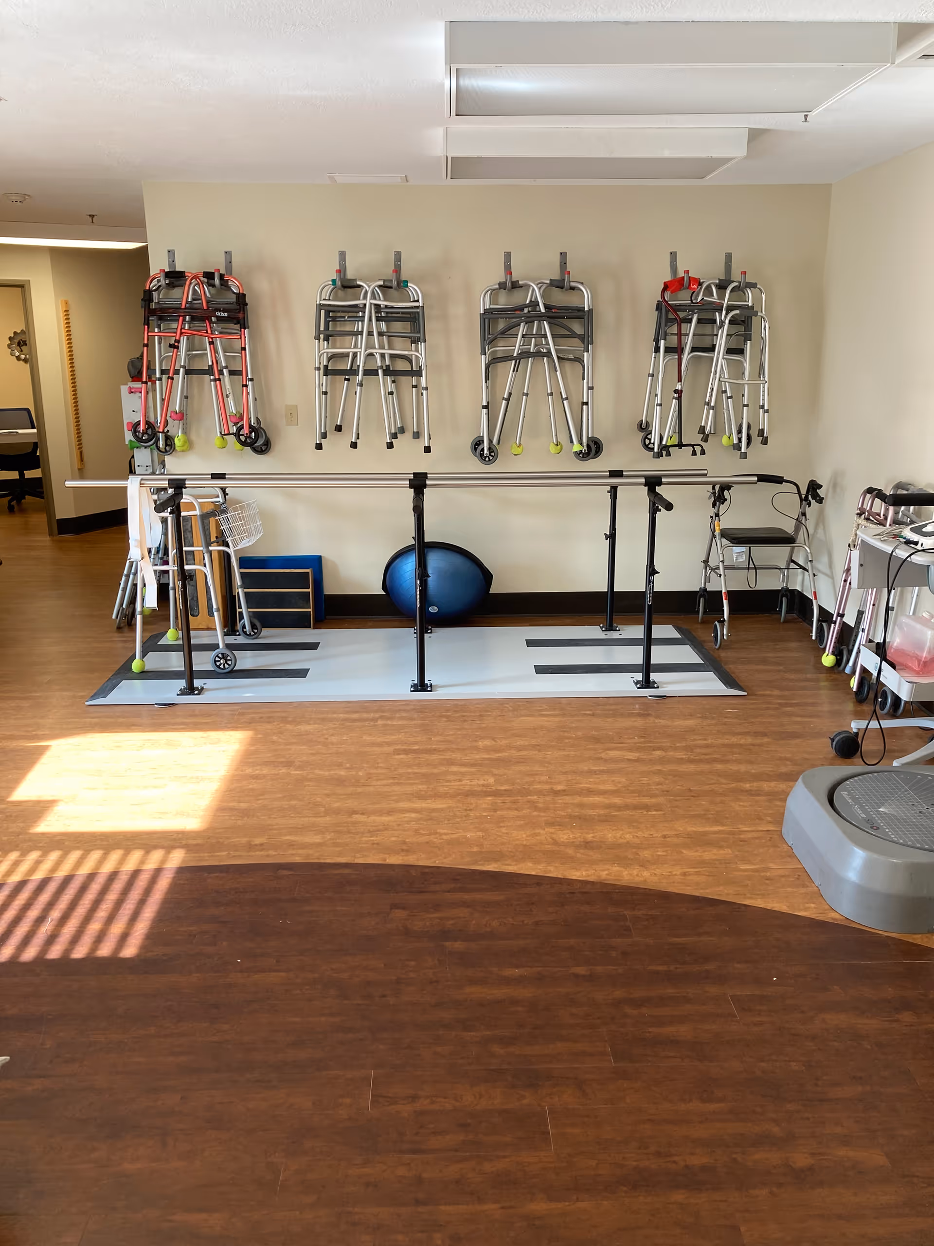 A rehabilitation or physical therapy room with multiple walkers hanging on the wall, parallel bars for walking exercises, a blue exercise ball, and other mobility aids on a wooden floor.