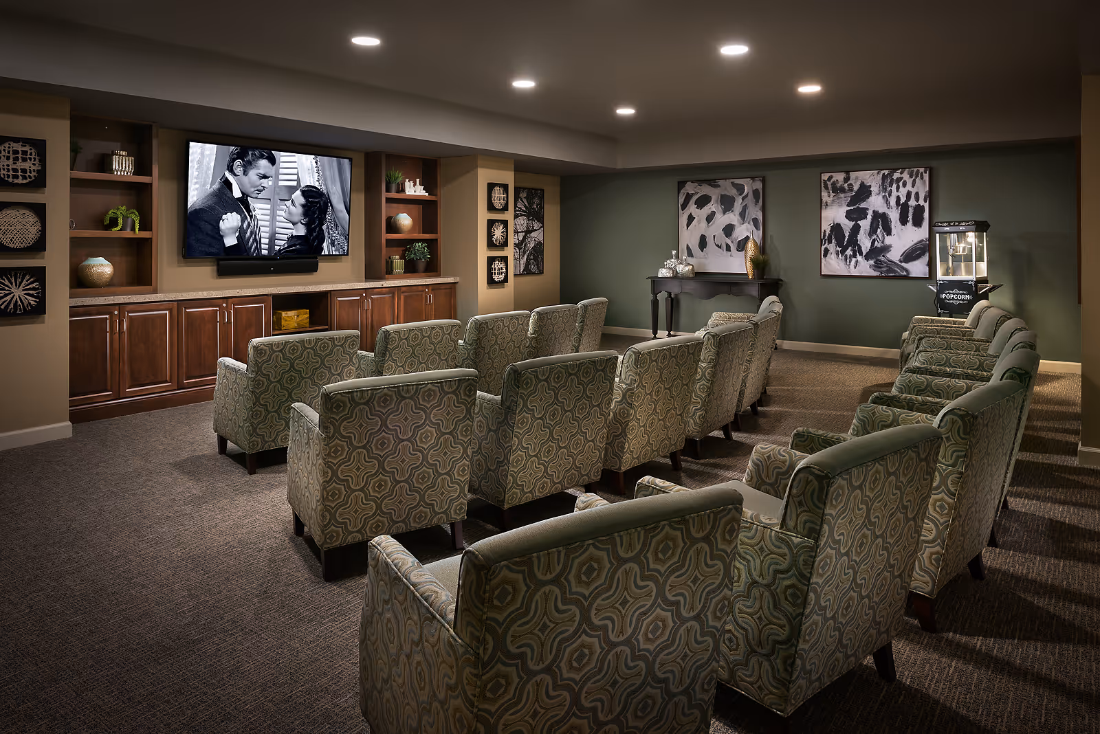 A cozy movie theater room with two rows of patterned armchairs facing a large flat-screen TV mounted on a wall with built-in wooden shelves and cabinets. The TV displays a black and white scene of a man and woman looking at each other. The room has soft lighting, green walls with abstract artwork, and a popcorn machine in the corner.