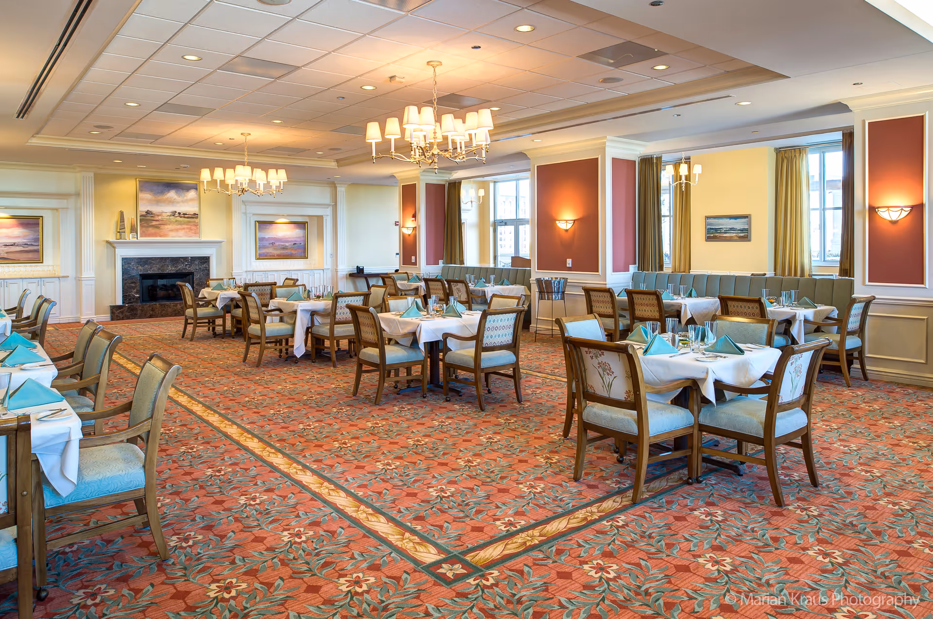 A spacious dining room with multiple tables covered in white tablecloths and set with turquoise napkins and glassware. The room features a patterned carpet, chandeliers, large windows with curtains, and framed artwork on the walls. There is a fireplace at the far end of the room.