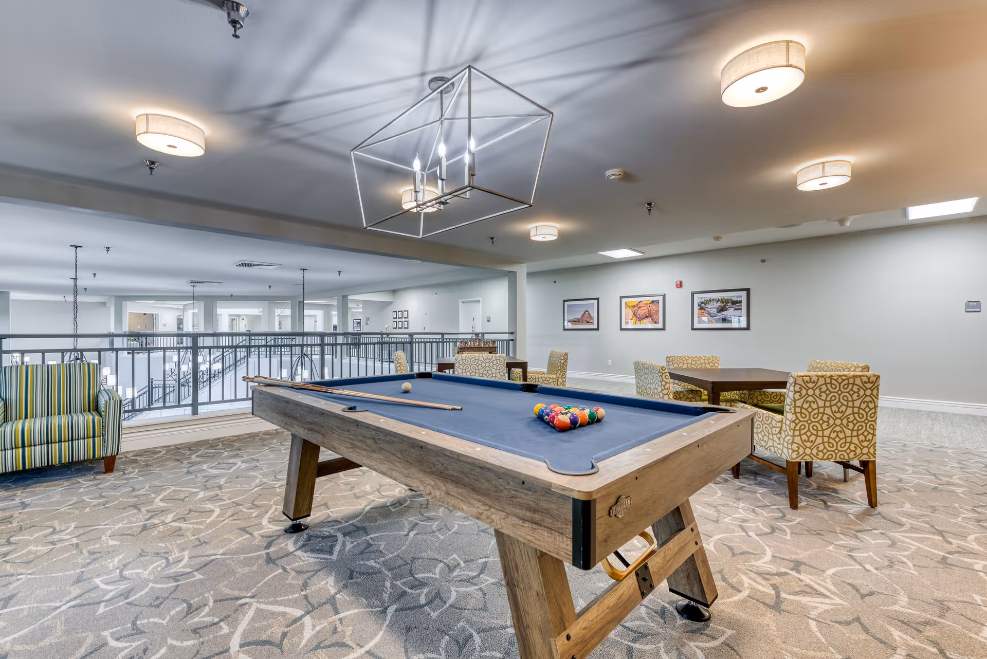 A spacious indoor recreational area with a pool table in the center, a striped armchair to the left, and two tables with patterned chairs in the background. The room has a patterned carpet, modern ceiling lights, and framed pictures on the far wall. A railing overlooks a lower level with more seating and lighting fixtures.