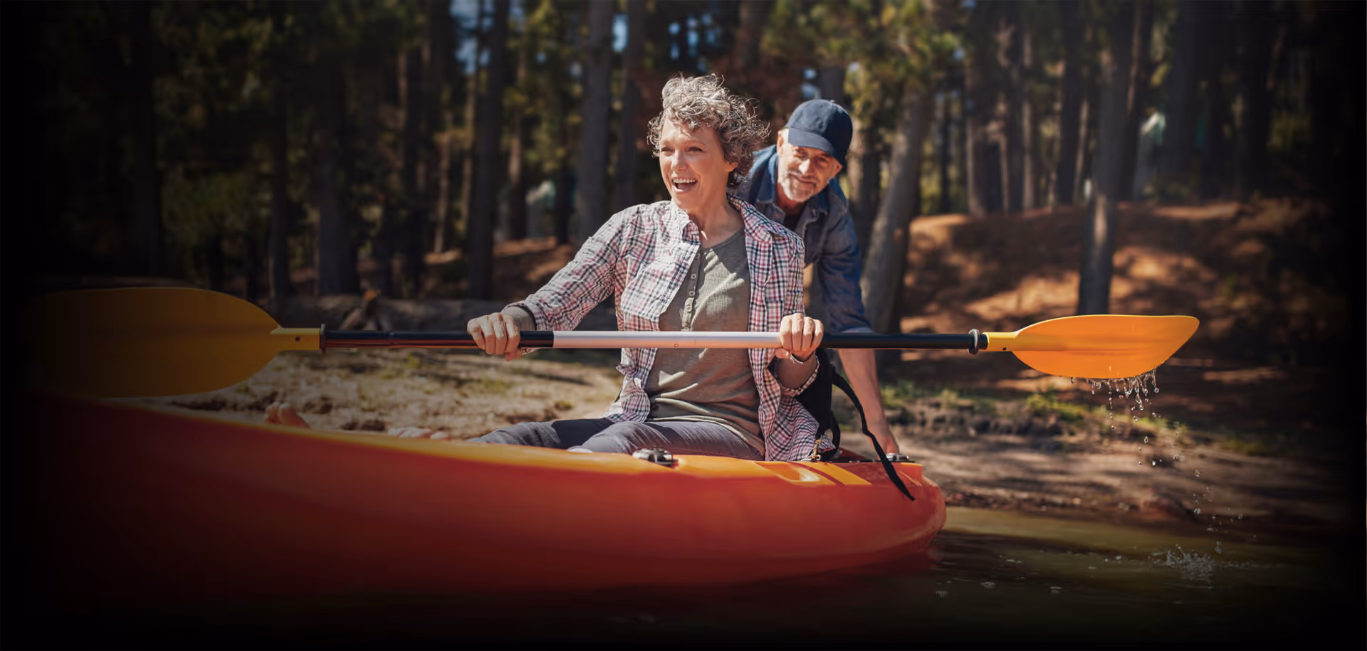 An elderly woman and man are kayaking together on a calm body of water surrounded by trees. The woman is sitting in front, smiling and holding a double-bladed paddle, while the man sits behind her, also holding the paddle.