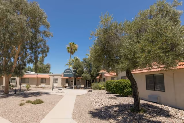 Outdoor view of Casas Adobes Post Acute Rehabilitation Center showing a paved walkway leading to the entrance with a blue canopy. The area is landscaped with trees, bushes, and gravel under a clear blue sky.