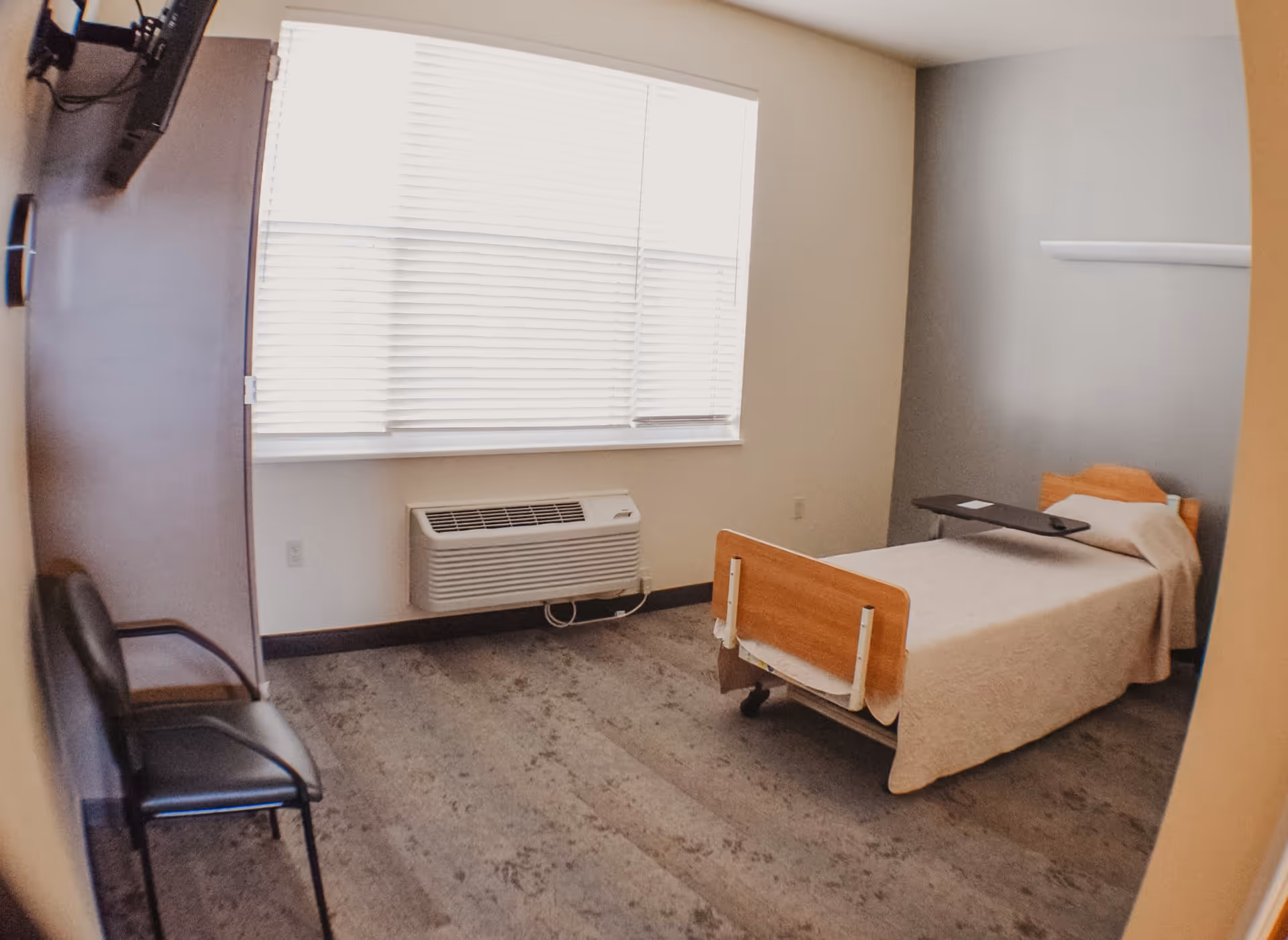 A simple, clean bedroom with a single hospital-style bed covered with a beige blanket, a black chair against the wall, a large window with white blinds, and a wall-mounted TV.