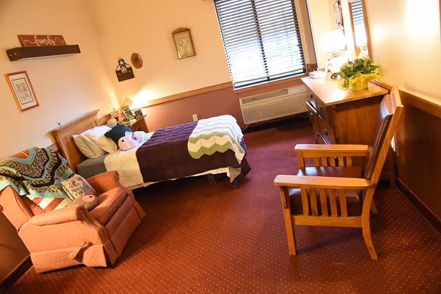 A cozy senior living bedroom with a single bed, upholstered armchair, wooden dresser and a window with blinds.