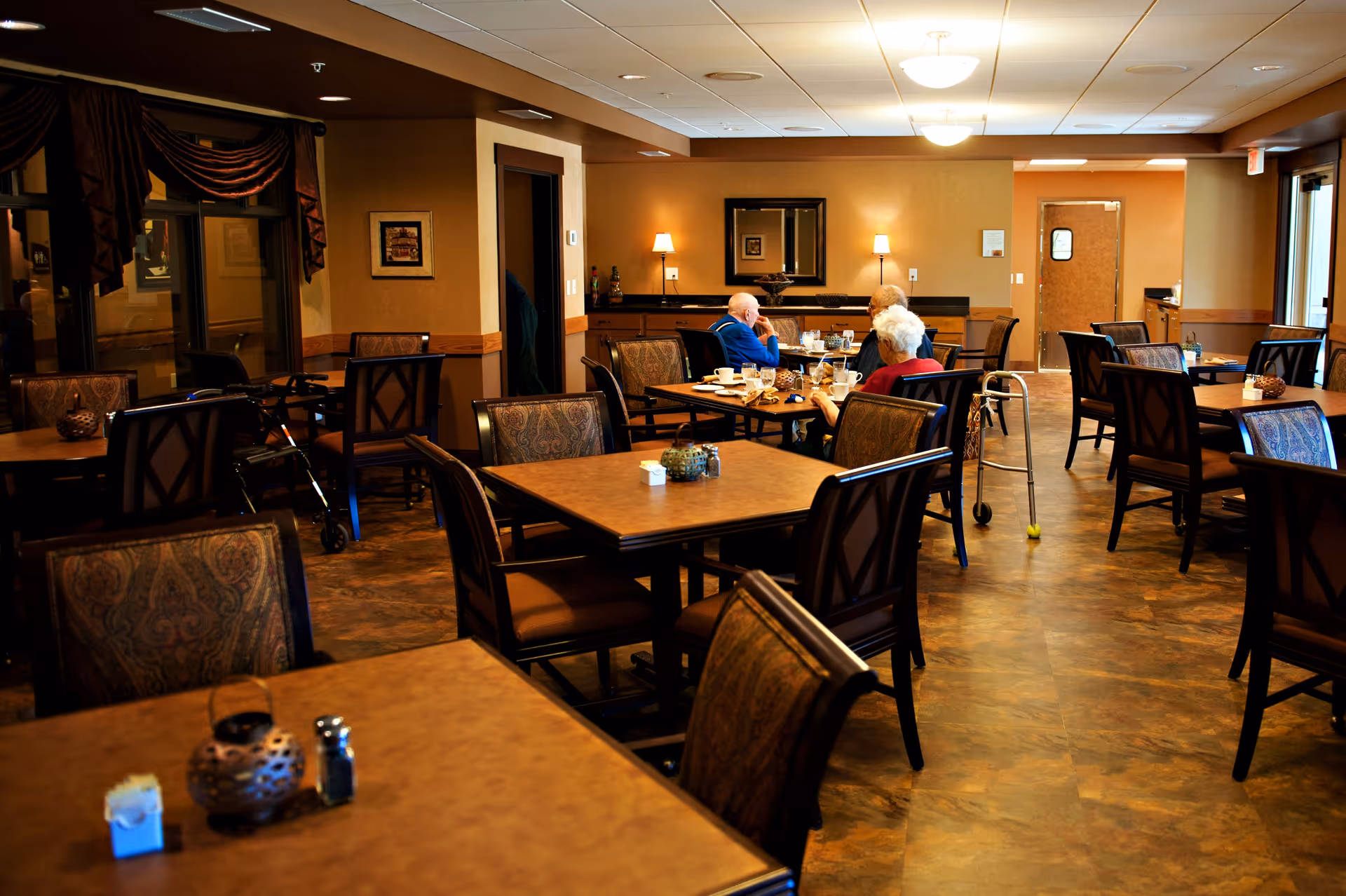 A dining room in an assisted living facility with several empty tables and chairs. Three elderly people are seated at one table, engaged in conversation. The room has warm lighting, brown walls, and a patterned floor. A walker is visible near the seated group, and there are decorative items on the tables.