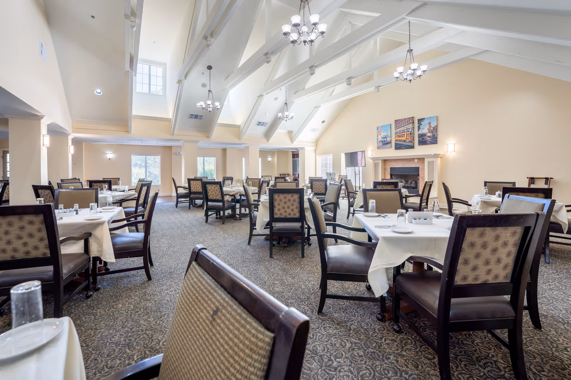 A spacious dining room with multiple tables covered in white tablecloths and set with plates, glasses, and utensils. The room features high vaulted ceilings with exposed white beams and several chandeliers. Large windows allow natural light to fill the space. There is a fireplace on one wall with three colorful paintings above it, and a television mounted nearby. The carpet has a patterned design, and the walls are painted a soft beige color.