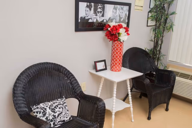 Two black wicker chairs with patterned cushions are placed on either side of a small white table. On the table is a red vase with red and white flowers and a small framed picture. A black and white framed photo of a woman is hanging on the wall above the table. A tall green plant is in the corner near a window with vertical blinds and an air conditioning unit below it.
