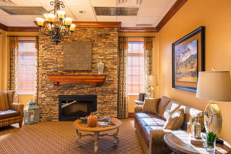 A cozy living room with a stone fireplace centered between two windows with wooden blinds and patterned curtains. The room features a brown leather sofa with decorative pillows, a round wooden coffee table with decorative items, a side table with a lamp and books, and a striped armchair. The walls are painted warm yellow, and a chandelier hangs from the ceiling. A framed landscape painting is on the wall above the sofa.