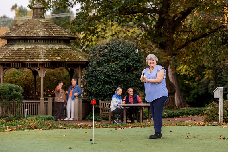 An elderly woman playing golf on a putting green in a garden area. In the background, there is a wooden gazebo with two elderly people standing and talking, and two more elderly people sitting at a table. The scene is surrounded by trees and greenery.