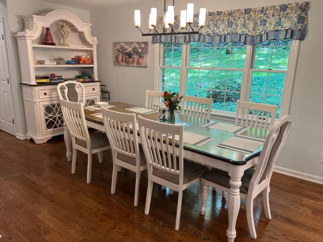 A bright dining room with a long wooden table surrounded by eight white chairs. The table is set with placemats and a small floral centerpiece. A large window with floral valance lets in natural light, showing green grass outside. A white hutch with shelves holding books and decorative items is against the wall, and a modern chandelier hangs above the table.