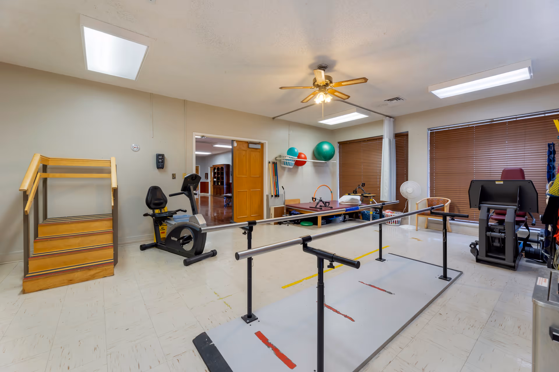 A rehabilitation therapy room with parallel bars for walking exercises, a stationary exercise bike, a small wooden staircase for step training, therapy balls on a shelf, a treatment table, and a leg press machine. The room has beige walls, a ceiling fan with lights, and windows with brown blinds.