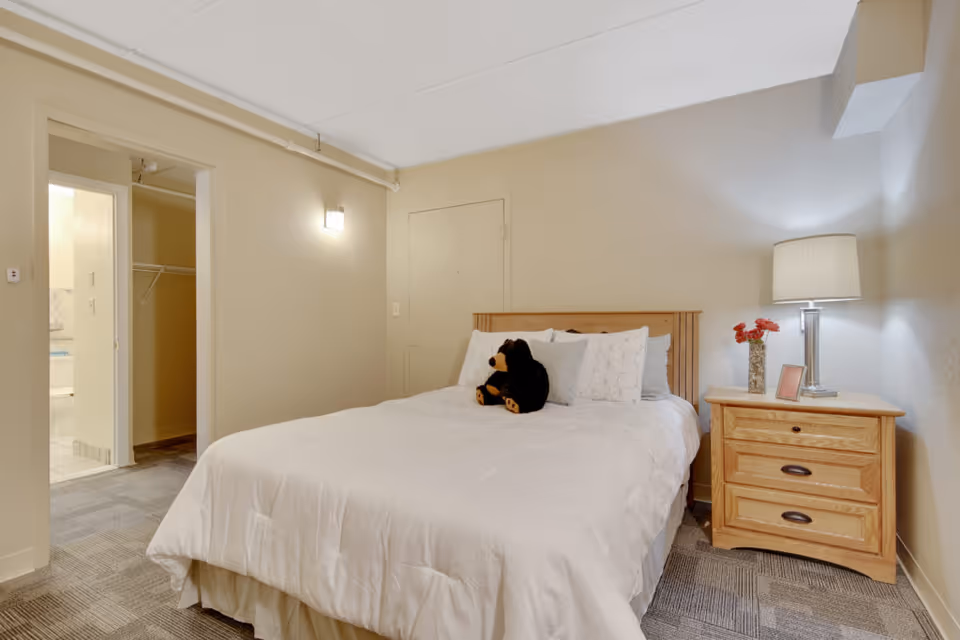Cozy bedroom with a made bed topped by a stuffed bear, a wooden nightstand with lamp and flowers, and an open doorway to a bathroom/closet.