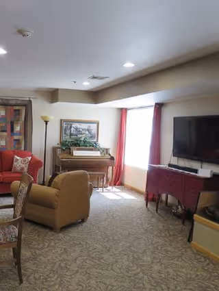 A cozy living room area with a beige carpet, a brown armchair, a red couch with patterned pillows, a wooden piano with a framed picture above it, a floor lamp, a window with red curtains, and a flat-screen TV mounted on the wall above a table covered with a red cloth.