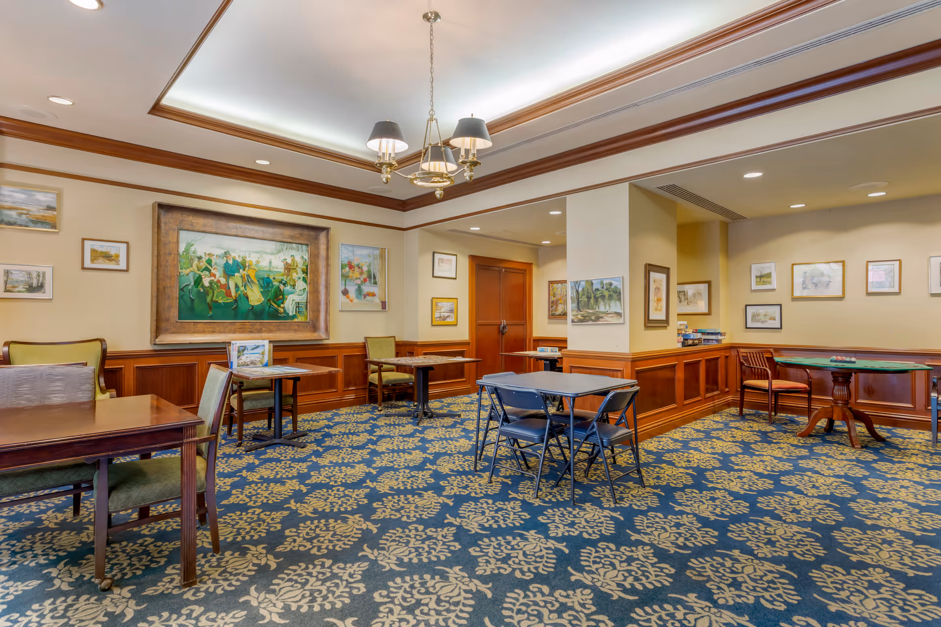 A spacious common area with multiple tables and chairs arranged for social or recreational activities. The room features a patterned carpet, wood-paneled walls with various framed paintings and artwork, and a ceiling light fixture with three lampshades. There is a card or game table with chairs in the corner and a stack of board games on a ledge.
