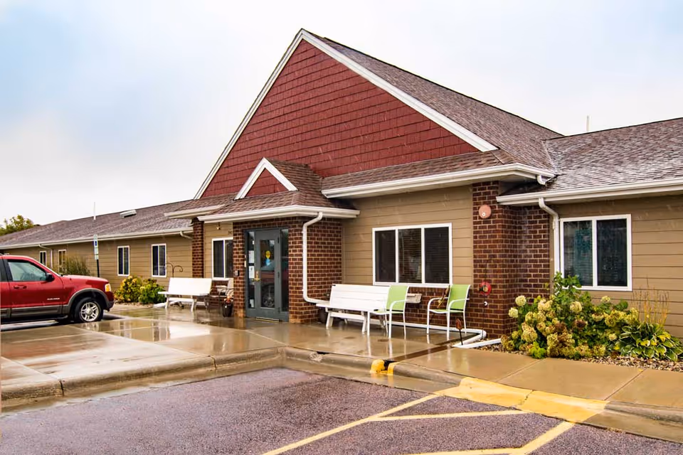 Exterior view of a single-story building with a red gable roof and beige siding. There are two white benches and two green chairs near the entrance door. A red vehicle is parked on the left side, and there are some plants and bushes near the building. The ground is wet, indicating recent rain.