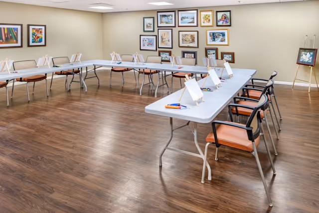 A spacious activity room with U-shaped tables and chairs arranged around them. Each table has small easels with blank canvases and paint supplies, suggesting a painting or art class setup. The walls are decorated with various framed artworks, and the floor has a wood finish.