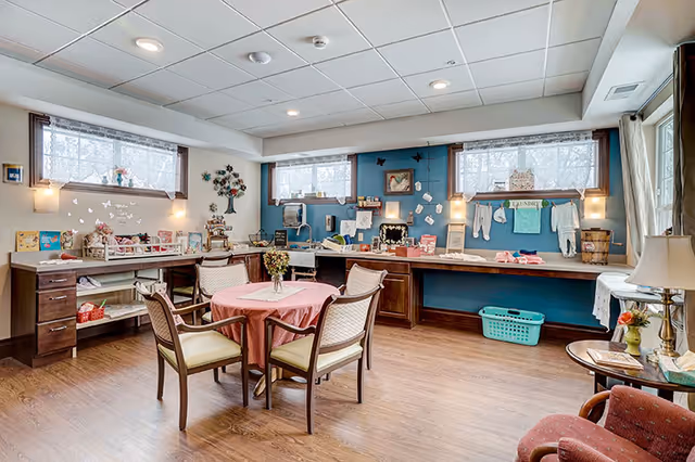 A bright and cozy activity room with a round table covered with a pink tablecloth and four chairs around it. The room has wooden flooring, large windows with lace curtains, and a long counter along the walls with various craft supplies, decorations, and laundry items. The walls are painted blue and decorated with butterflies and a tree wall art. There is a comfortable armchair and a small side table with a lamp and flowers in the corner.